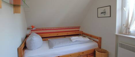 Un petit lit en bois avec un seul oreiller et une couverture, un panier en osier et un tableau encadré accroché au mur.