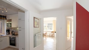 Een moderne keuken met witte kasten, een ingebouwde oven en een koffiezetapparaat. Een eethoek met een tafel en stoelen is zichtbaar door een open deur.