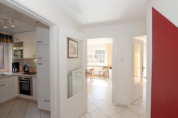 A modern kitchen with white cabinetry, a built-in oven, and a coffee maker. There is a dining area with a table and chairs visible through an open door.