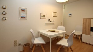 A dining area with a white table and chairs, a wooden cabinet, and a wall-mounted shelf with framed pictures and clocks. - Ferienwohnung mit 2-zimmern mit ca. 45m² und Balkon (Wyk auf Föhr)