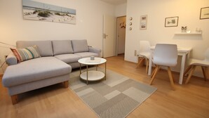 A modern living room with a grey sofa, a round coffee table, and a dining area with a white table and chairs. - Ferienwohnung mit 2-zimmern mit ca. 45m² und Balkon (Wyk auf Föhr)
