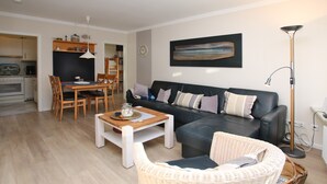 A modern living room with a black sofa, a dining table, and a wicker chair. - Ferienwohnung in Wyk/ Südstrand mit ca. 56m² und 3- Zimmern in der Ersten Etage (Wyk auf Föhr)