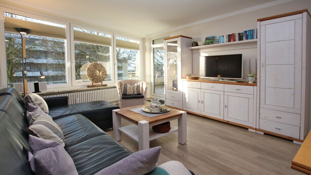 A modern living room with a sofa, a coffee table, and a television. There is a window with a view of trees and a radiator under the window. - Ferienwohnung in Wyk/ Südstrand mit ca. 56m² und 3- Zimmern in der Ersten Etage (Wyk auf Föhr)
