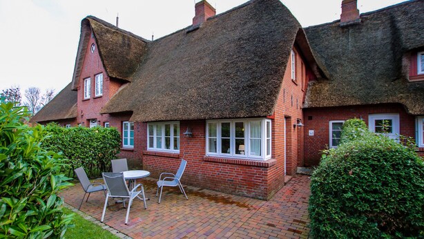 A red brick house with a thatched roof and a small patio with a table and chairs. - Ferienwohnung in Wrixum, mit 2- Zimmern auf 51m² im Reetdachhaus (Wrixum)