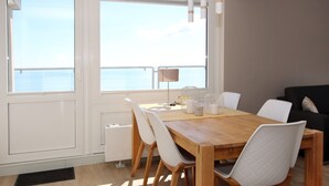 Un comedor con mesa de madera y sillas blancas, ventanas amplias con vista al mar y un sofá negro al fondo.