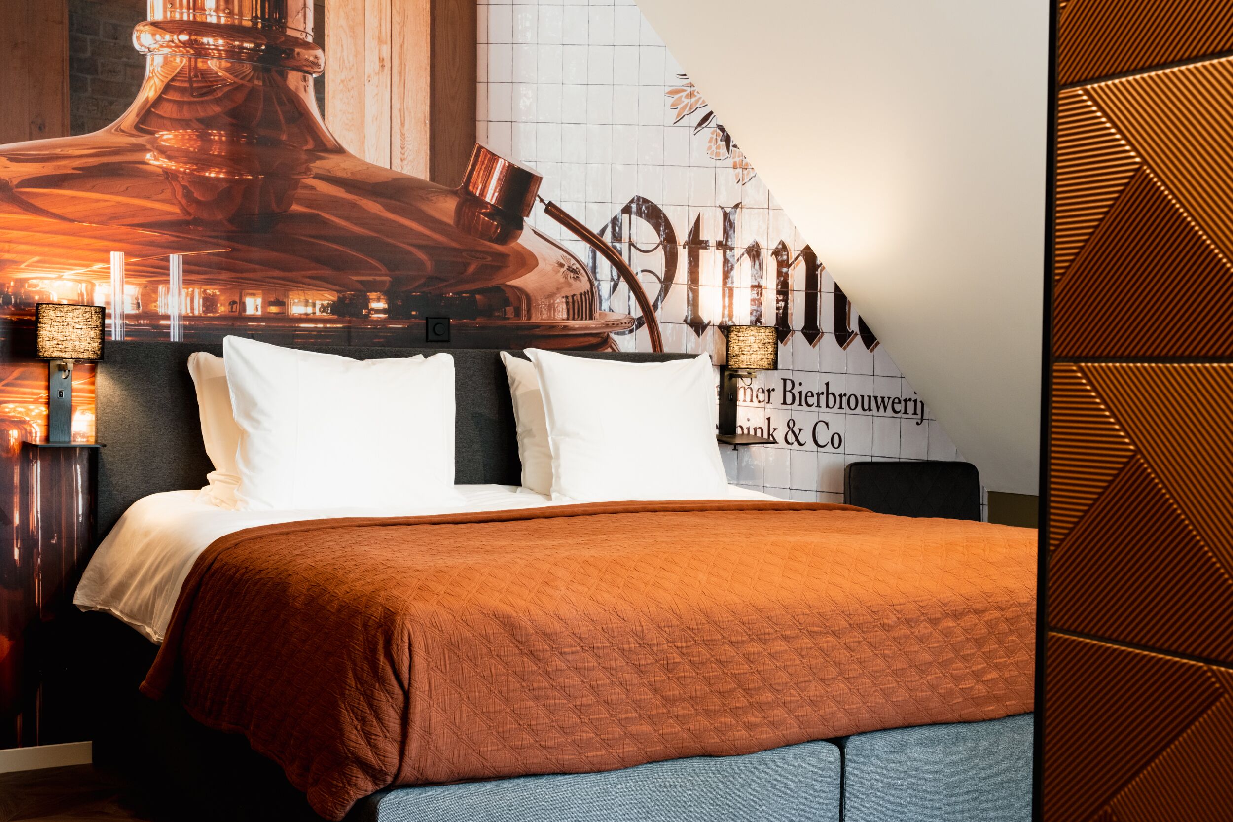 A hotel room with a large copper distillation apparatus as a wall decoration, a bed with white and brown bedding, and a sign that reads "Bierbrouwerij Pink & Co."
