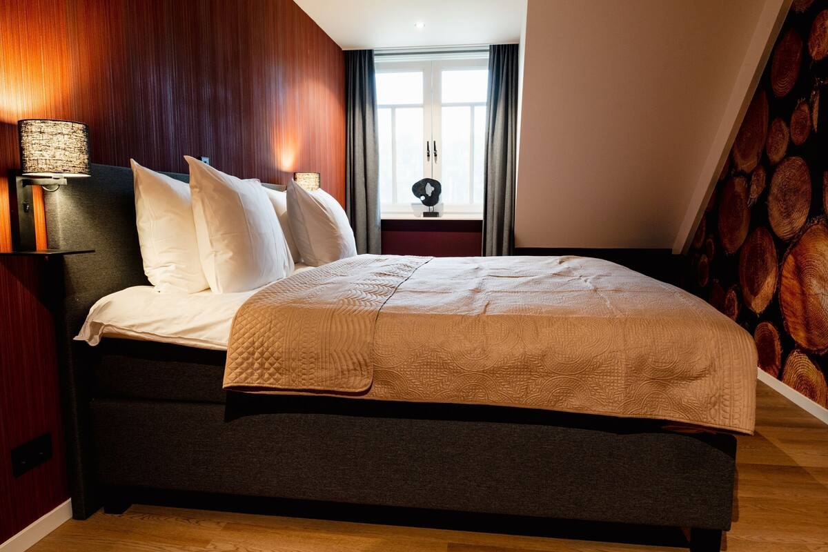 Une chambre à coucher comprenant un lit, une table de chevet, une lampe, une fenêtre avec des rideaux et un mur au motif de bois.