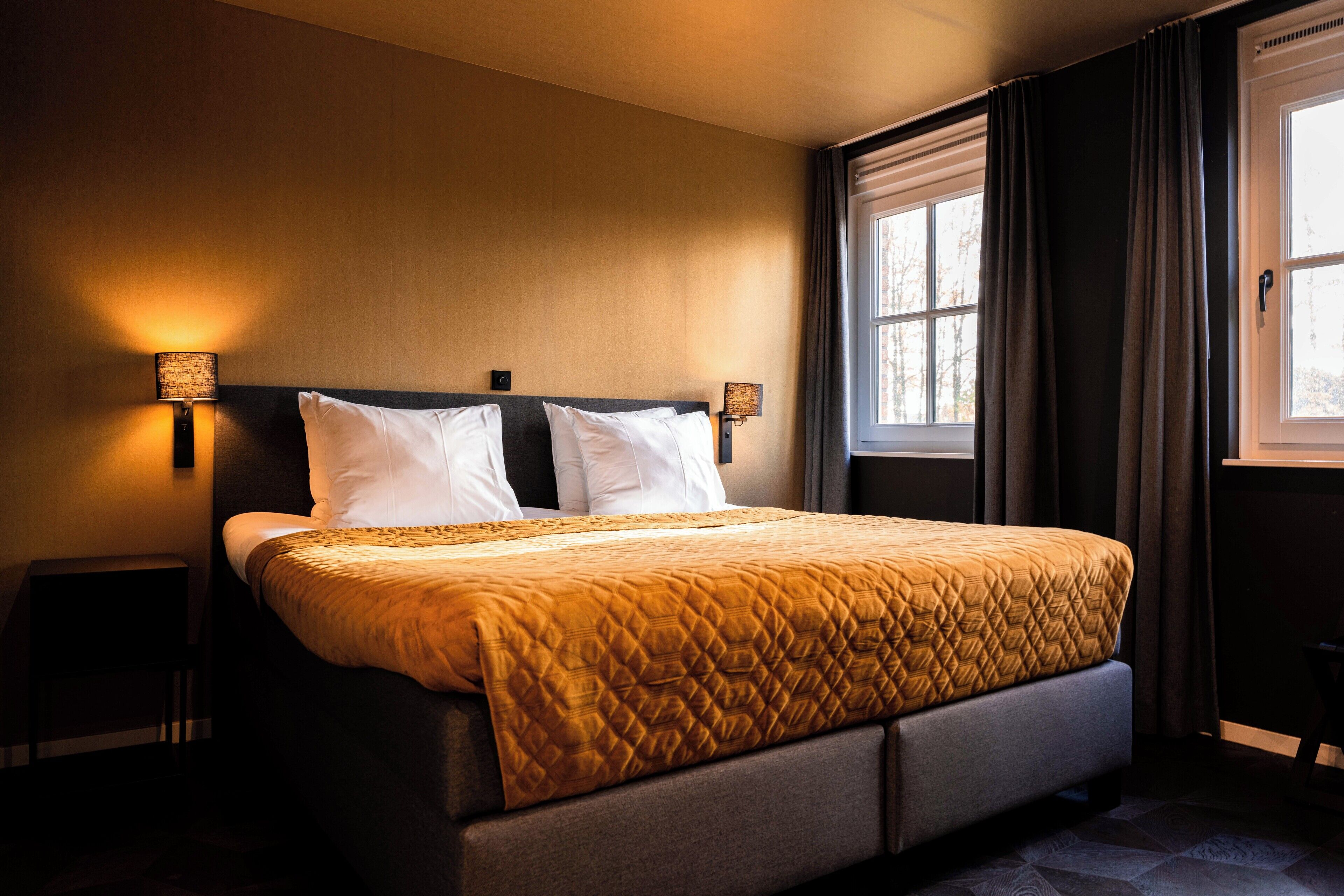 A neatly made bed with a quilted cover, two bedside lamps, and a window with curtains.
