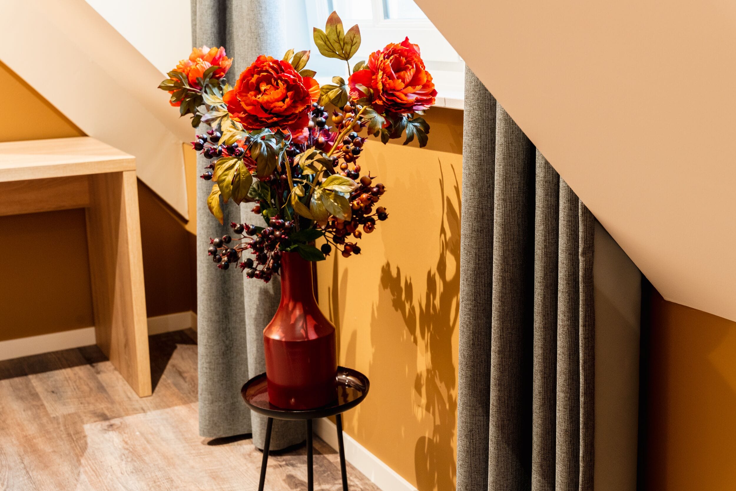 Un jarrón rojo con flores sobre una mesa pequeña en una habitación con suelo de madera y una escalera.