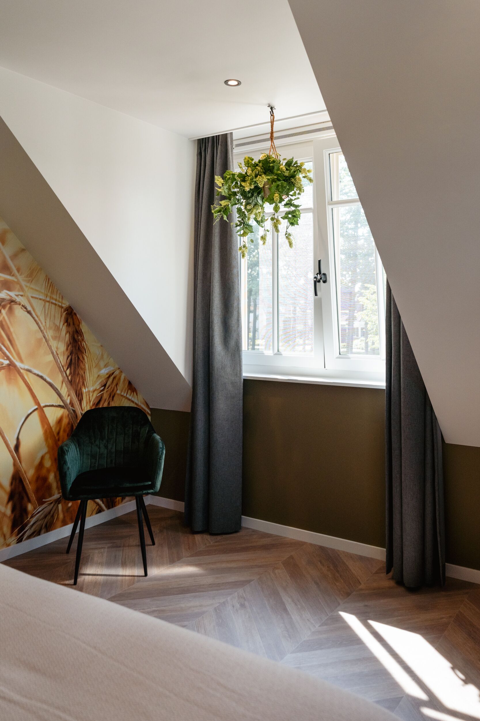 A room with a sloped ceiling, a window with curtains, a hanging plant, a chair, and a wall with a wheat pattern.