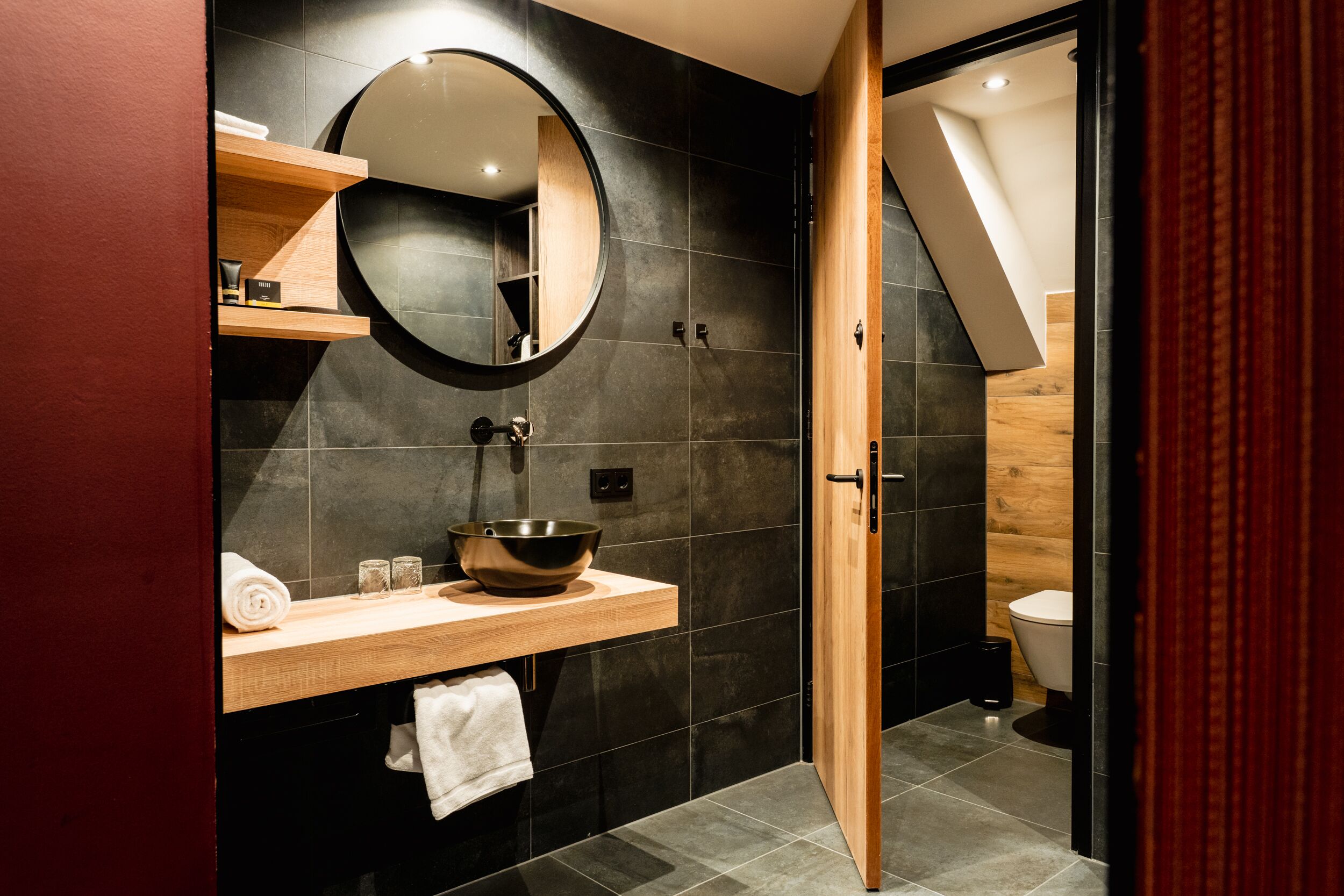 Een moderne badkamer met een houten wastafelmeubel, een ronde spiegel en een wand betegeld met zwart tegelwerk.