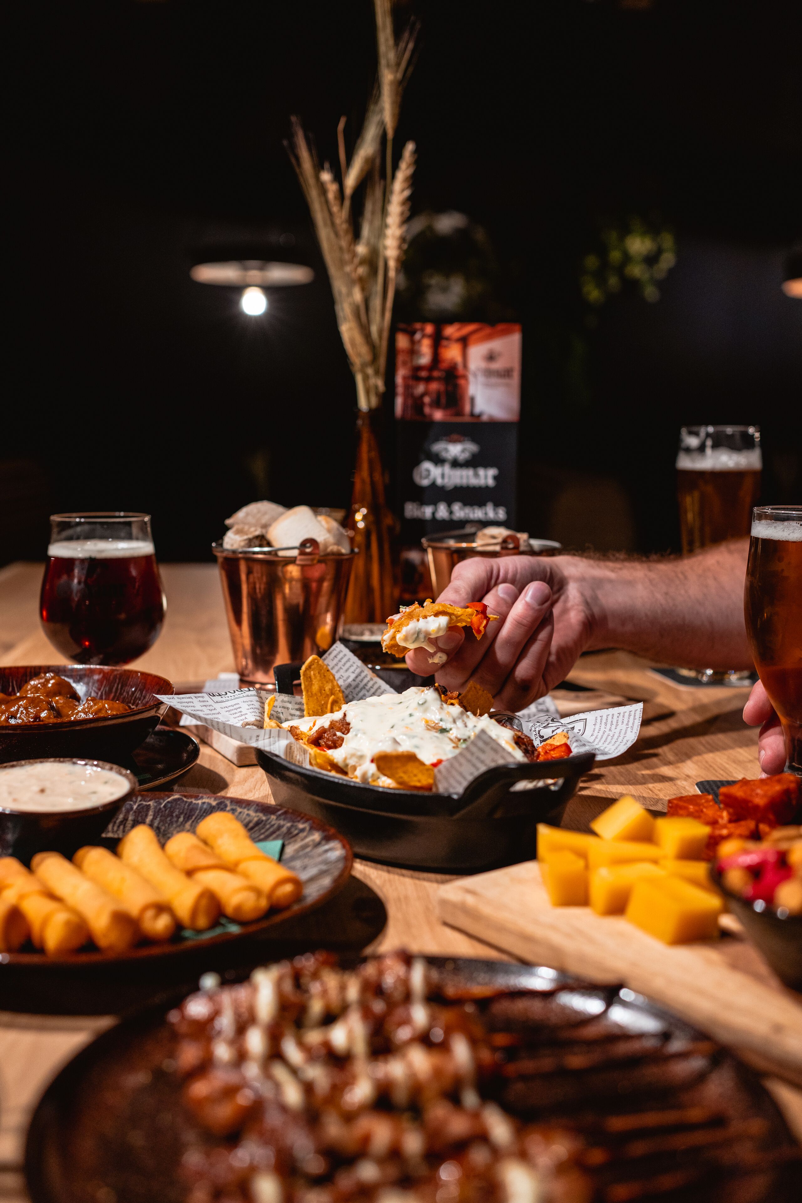 Une table à manger avec divers aliments et boissons, notamment une chope de bière, un verre de bière et une assiette de nachos.