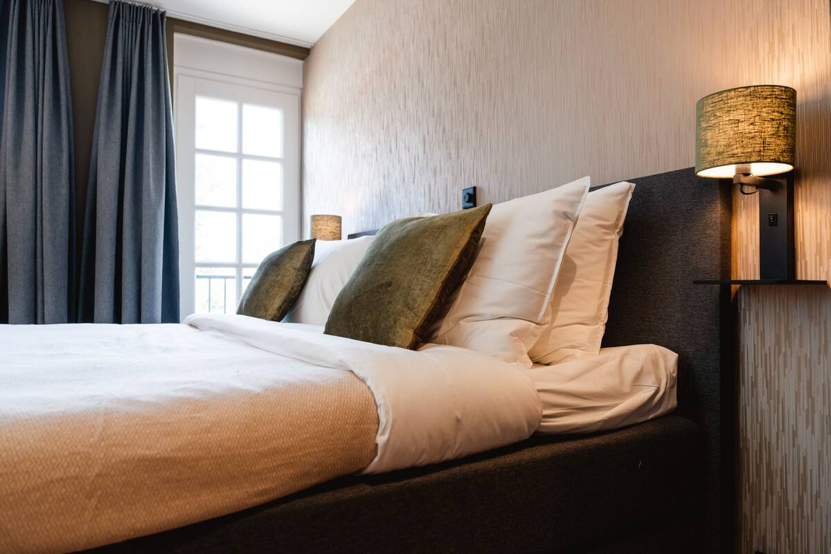 Un lit bien fait, avec du linge de lit blanc et plusieurs oreillers, une lampe fixée au mur et une fenêtre avec des rideaux.