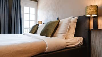 Cama bem arrumada com lençóis brancos e várias almofadas, uma luminária na parede e uma janela com cortinas.
