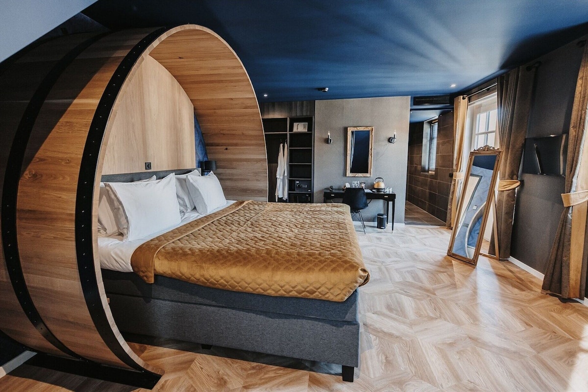 Une chambre à coucher dotée d’une tête de lit originale en forme de tonneau, d’un lit recouvert d’une housse matelassée, d’un bureau avec une chaise et d’un miroir accroché au mur.