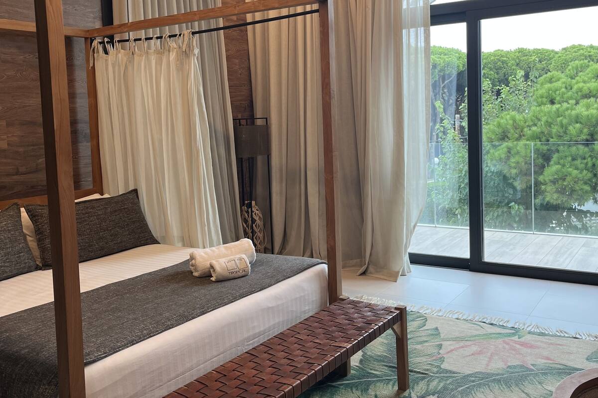Een slaapkamer met een hemelbed, een bankje en uitzicht op groen door grote ramen.