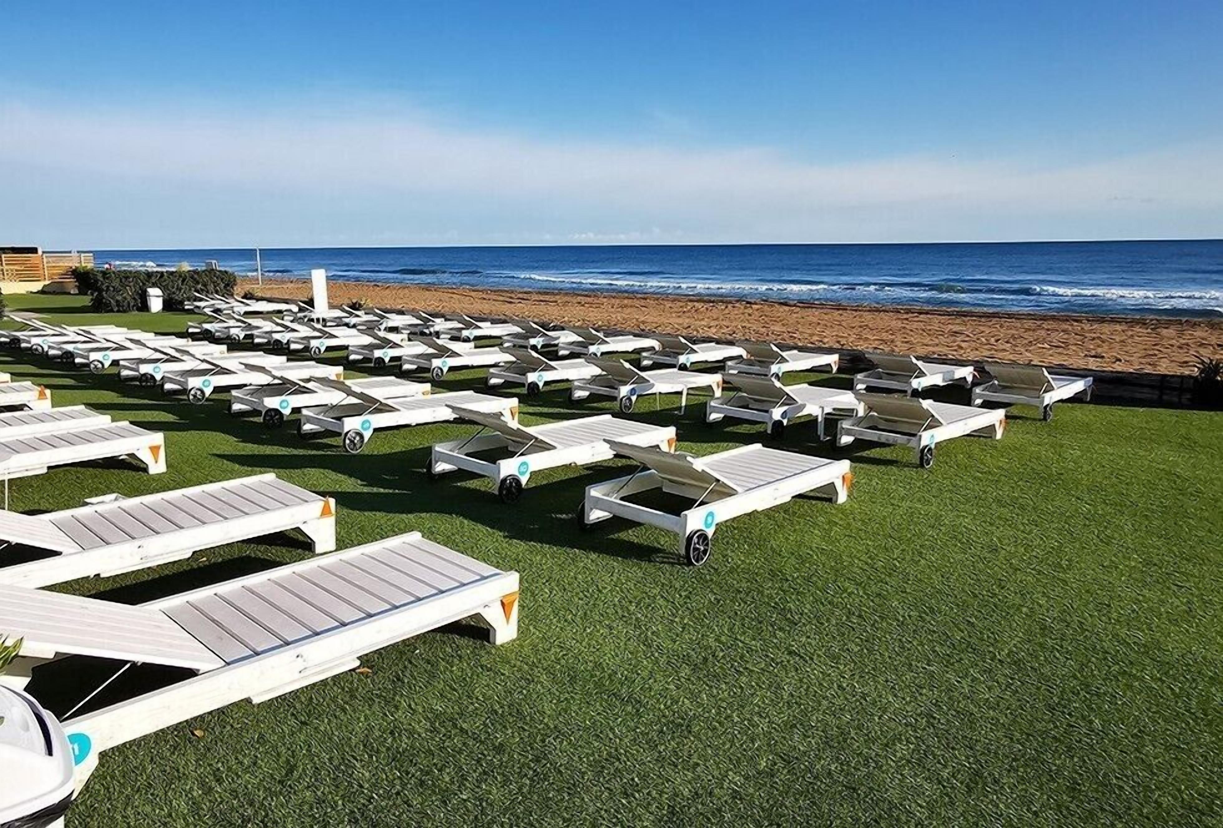 A beachside lounge area with rows of white lounge chairs on artificial grass, facing the ocean.