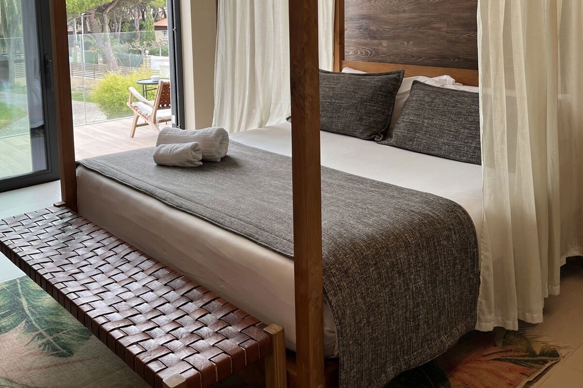A four-poster bed with a bench, a large window, and a view of the outdoors.