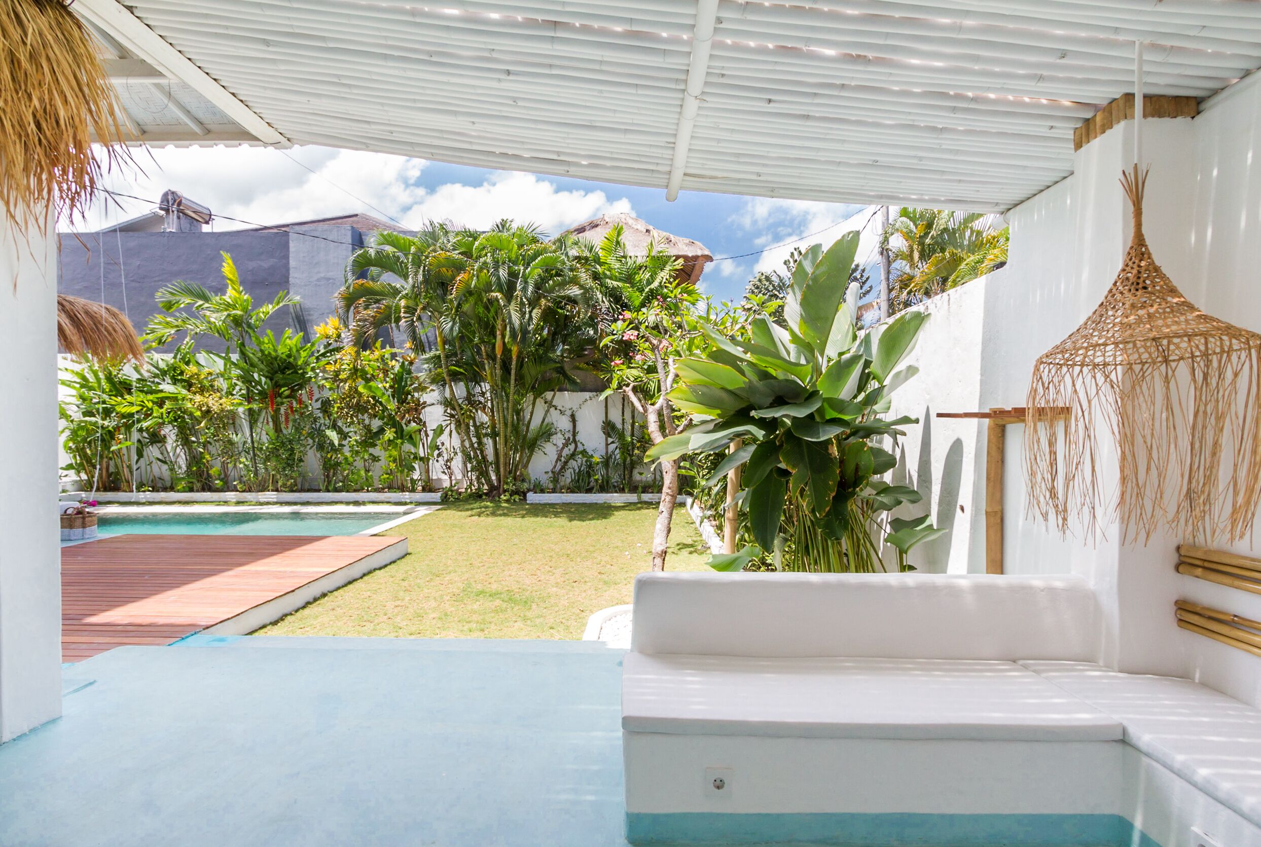 A pool area with a wooden deck, a white bench, and lush tropical plants.