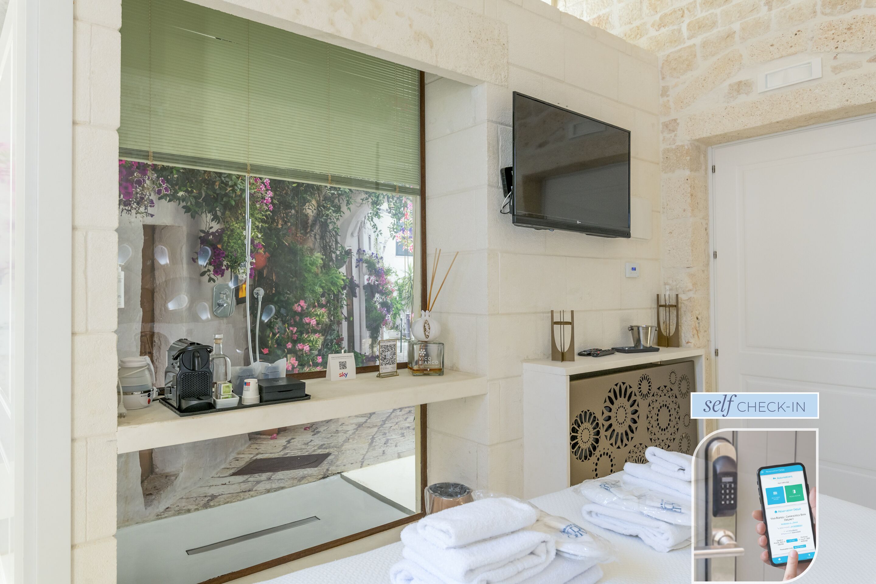 A hotel room with a television mounted on the wall, a window with a view of greenery, a self-check-in kiosk, and neatly folded towels on the counter.