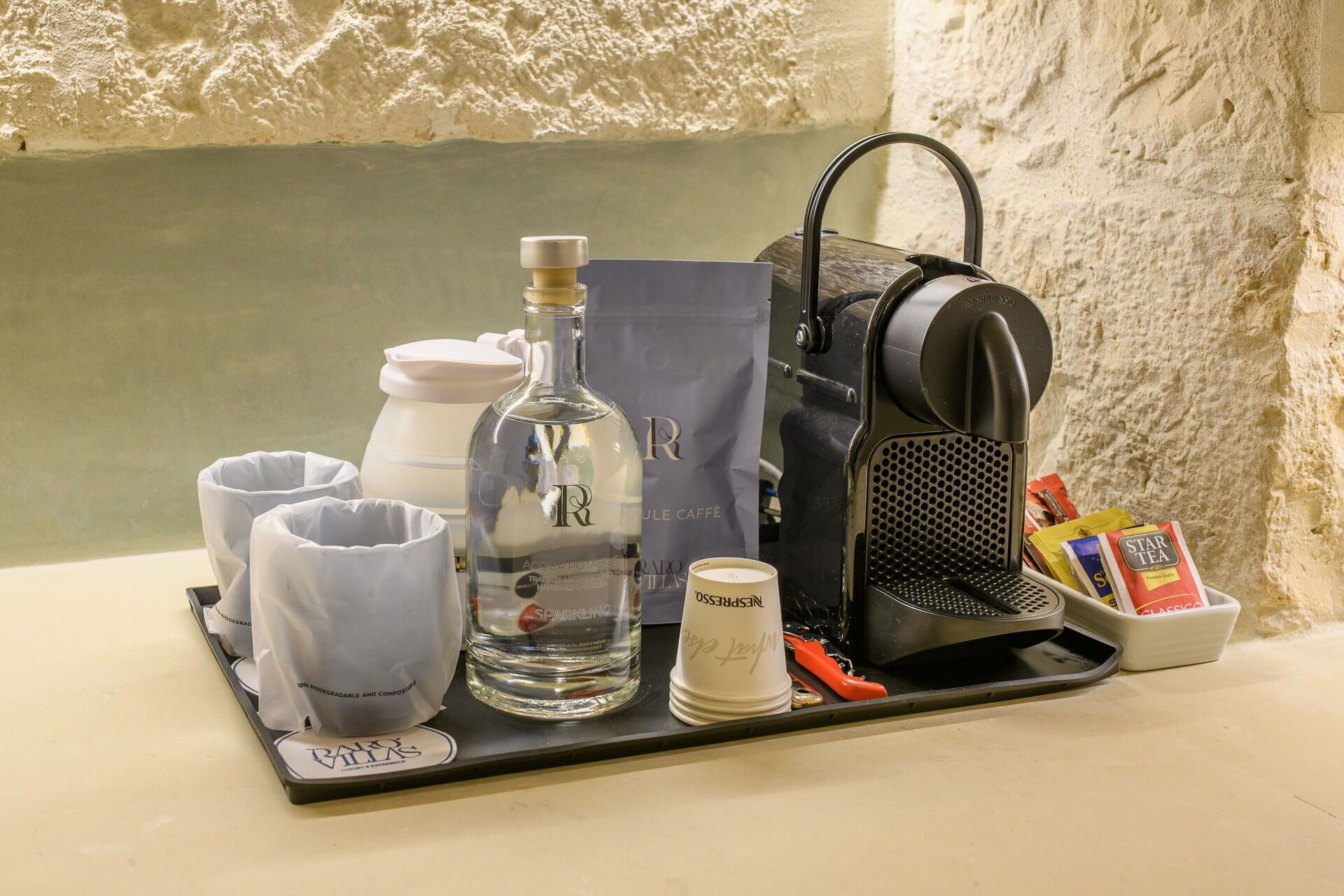 A coffee station with a coffee machine, cups, a bottle of water, a bag, and some tea packets.