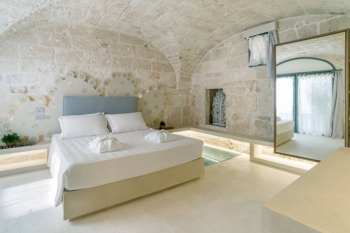 A bedroom with a stone wall, a bed with white linens, a nightstand, and a window with a view.