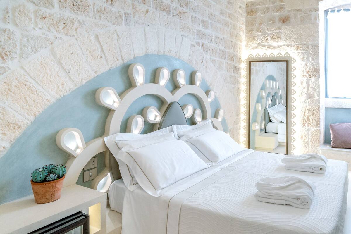 A bedroom with a stone wall, a bed with white linens, a potted plant, and a mirror.