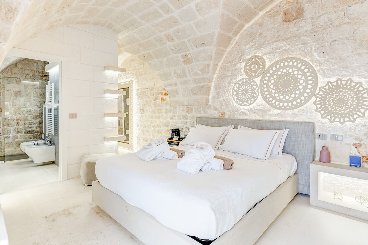 A spacious bedroom with a large bed, a bedside table with a lamp, and a stone wall.