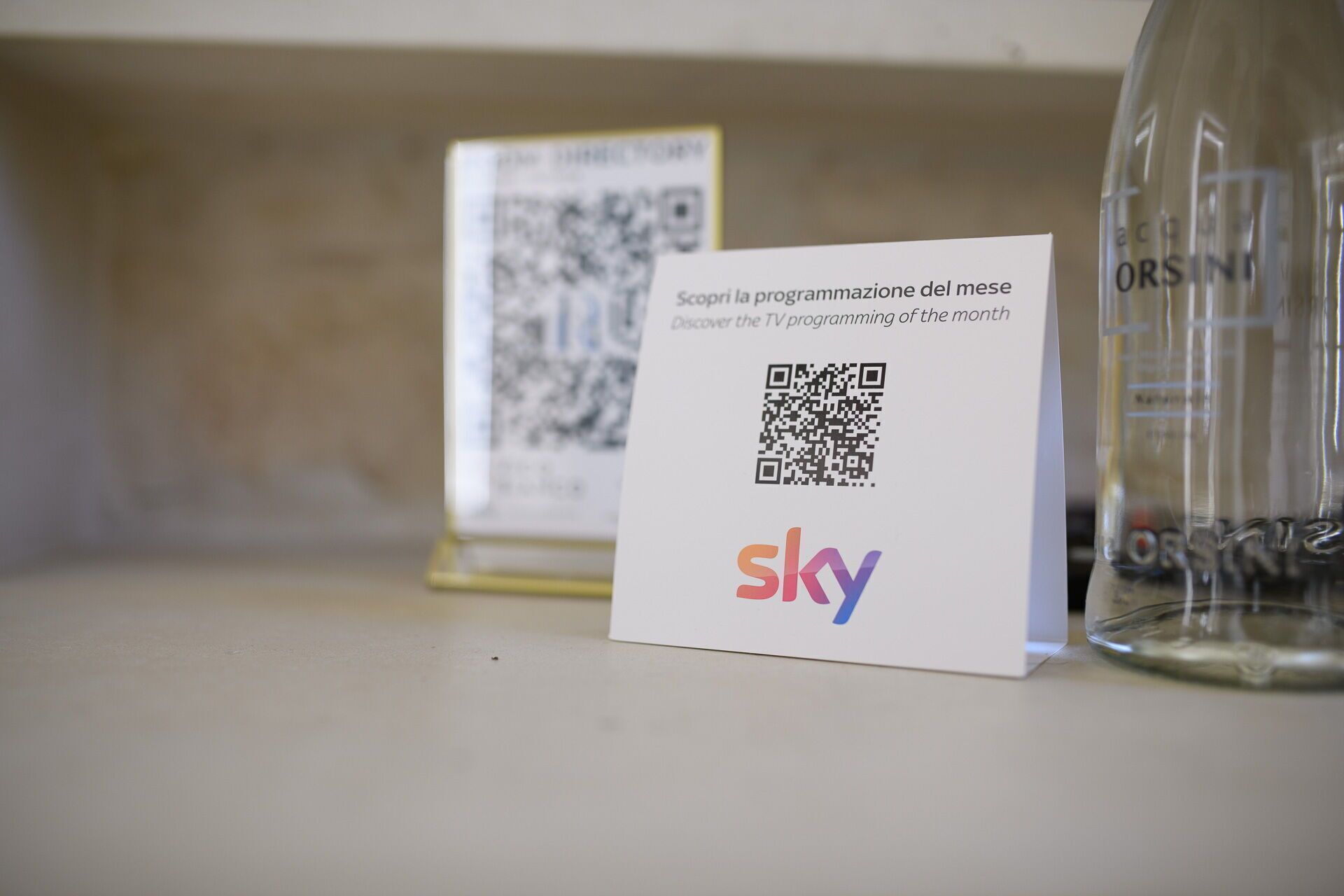 A QR code on a card with the Sky logo, placed on a shelf next to a clear bottle.