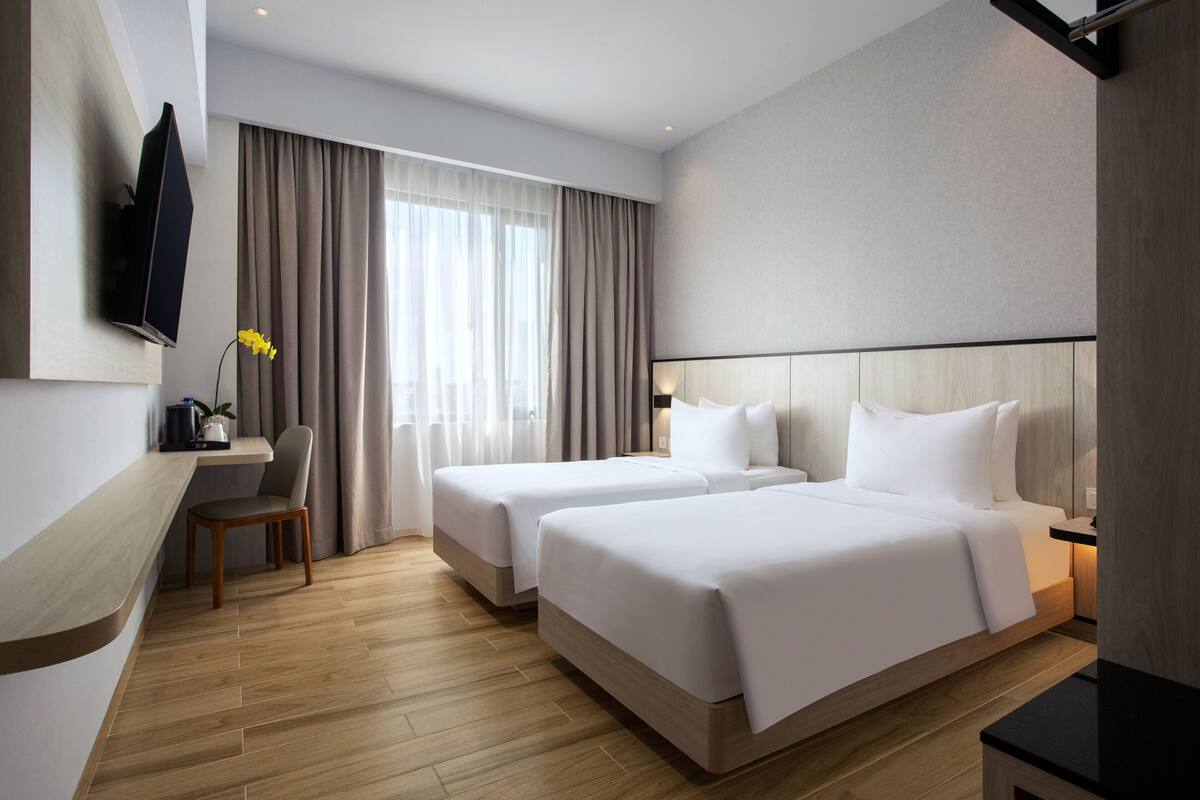 A hotel room with two beds, a TV mounted on the wall, a desk with a chair, and a large window with curtains.