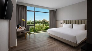 Habitación de hotel con una cama grande, un escritorio con silla, un televisor de pantalla plana y vistas a un paisaje urbano a través de amplios ventanales.