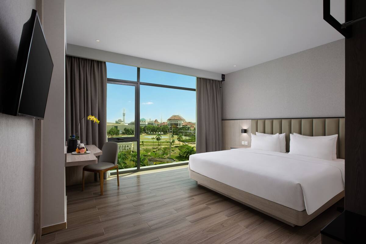 A hotel room with a large bed, a desk with a chair, a flat-screen TV, and a view of a cityscape through large windows.