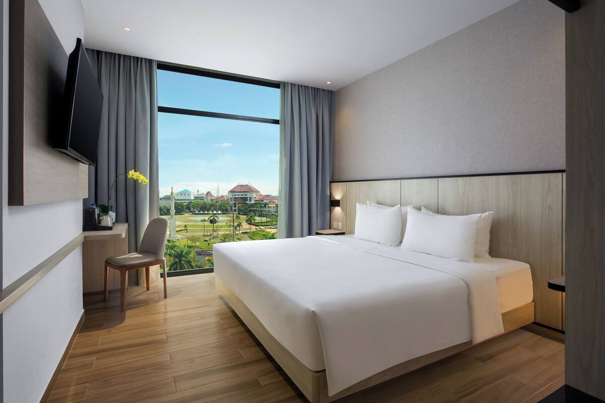 A modern hotel room with a large bed, a TV mounted on the wall, a desk with a chair, and a view of a cityscape through large windows.