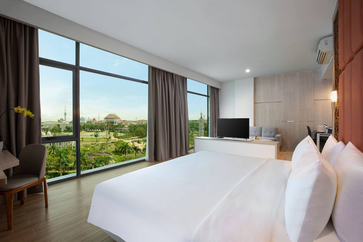 A modern hotel room with a large bed, a TV, a desk, and a view of a cityscape and greenery.