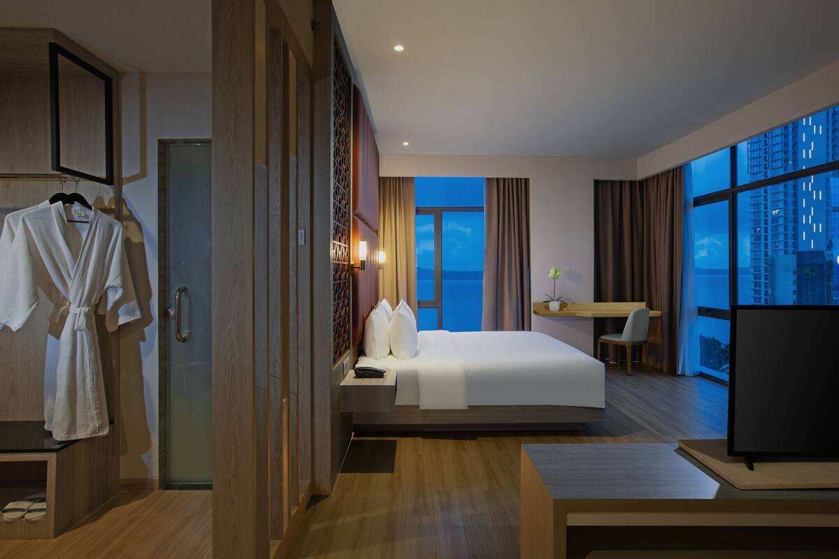 A hotel room with a bed, a desk, a TV, and a view of the city at night.