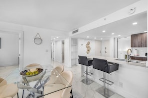 A modern kitchen with a central island, bar stools, and a dining area with a glass table and chairs. - Chic Bayfront Condo With Stunning View (Miami)