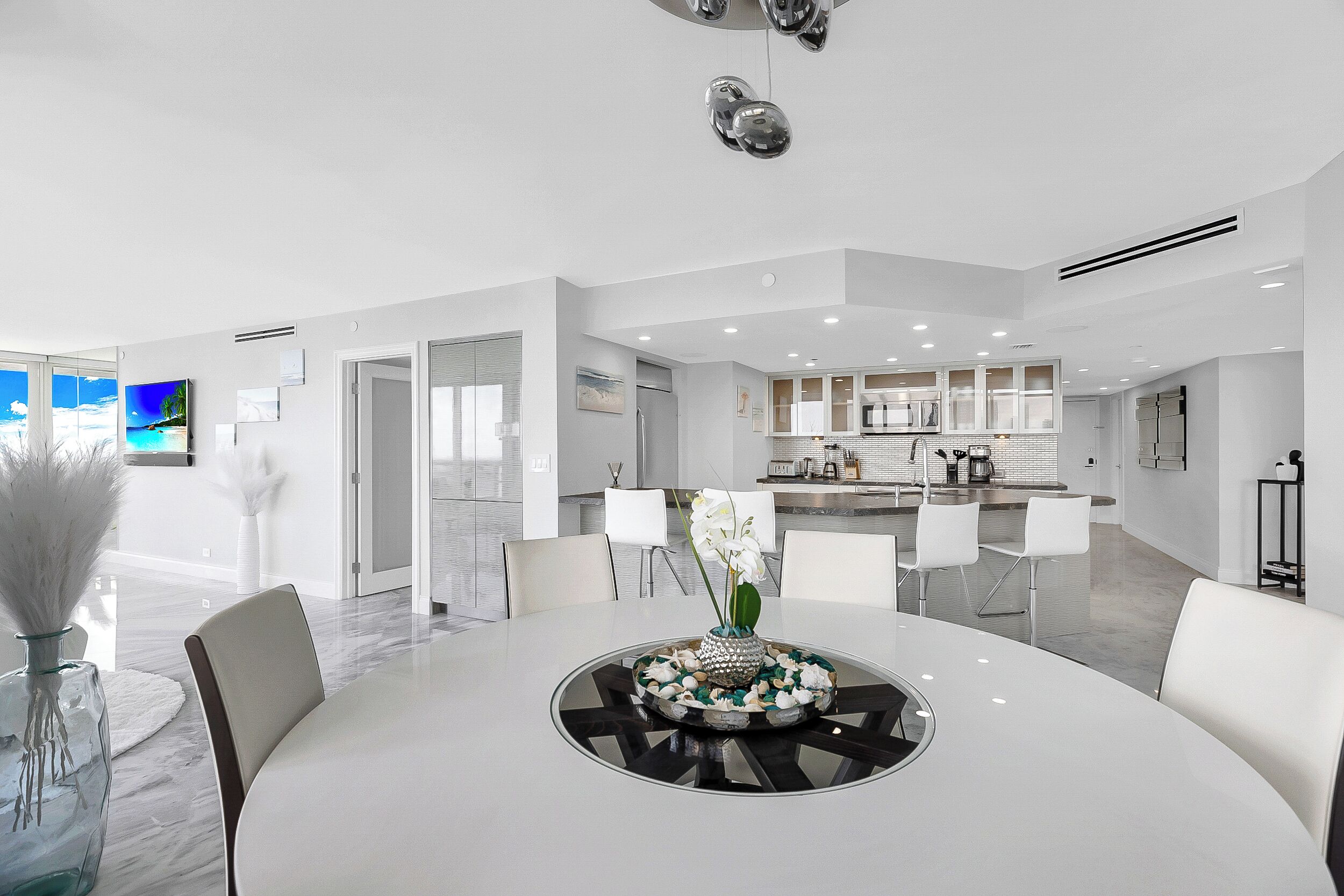 A modern, spacious living area with a central dining table, white chairs, and a kitchen in the background.