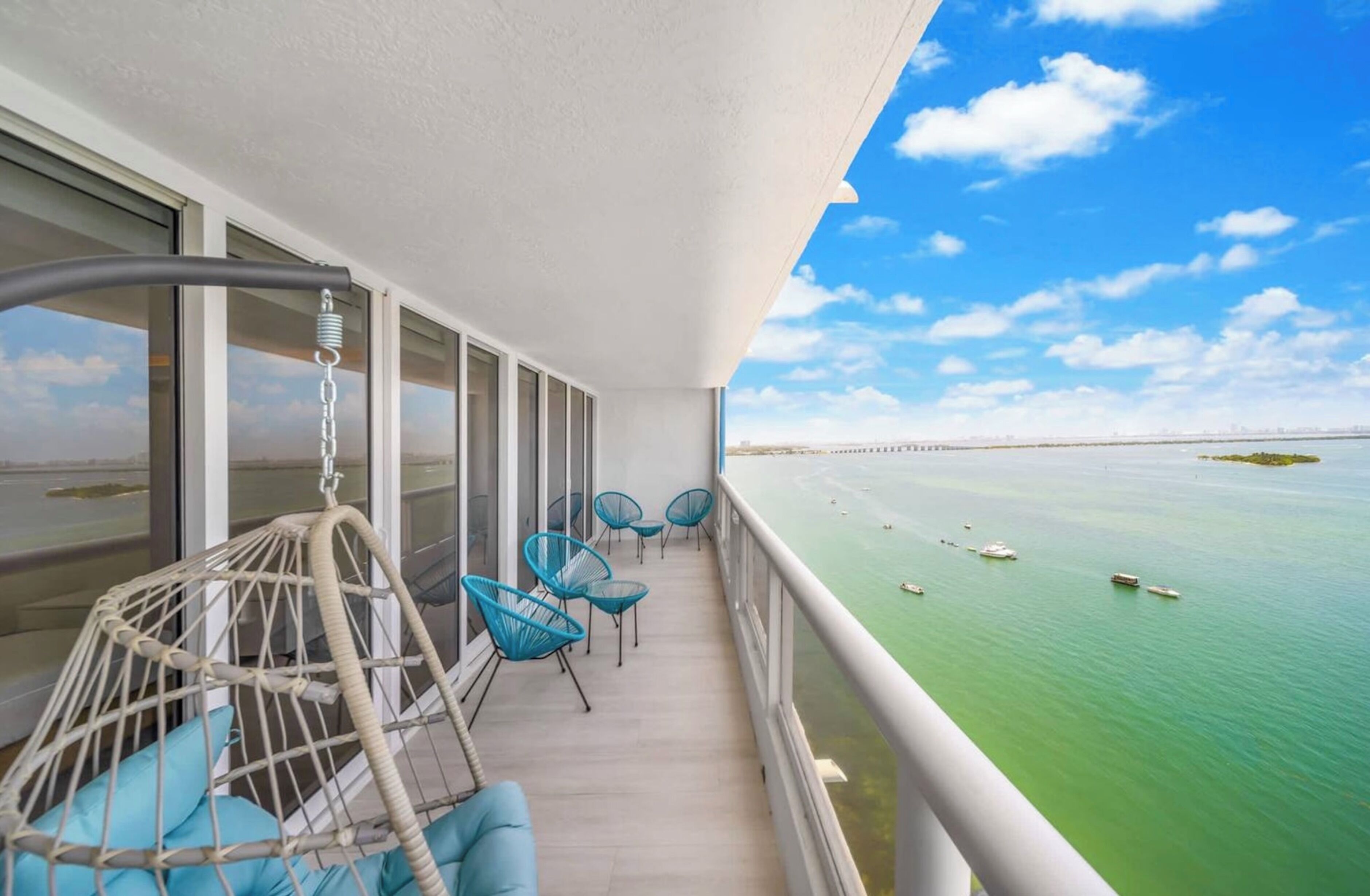 Ein Balkon mit Korbstühlen und einer Hängematte, mit Blick auf ein ruhiges Meer und Boote.