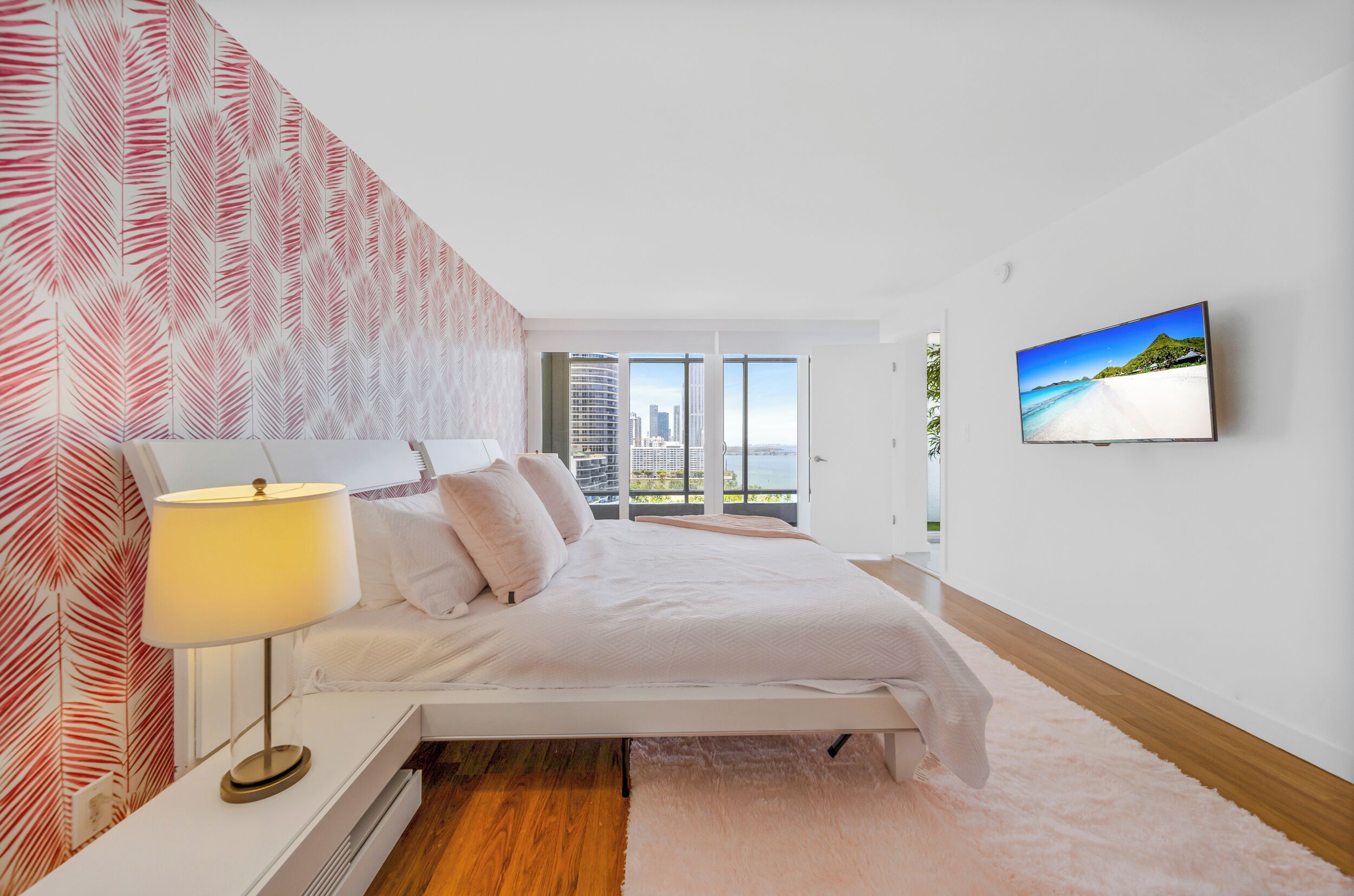 A modern bedroom with a bed, a bedside table with a lamp, a large window with a city view, and a TV on the wall.
