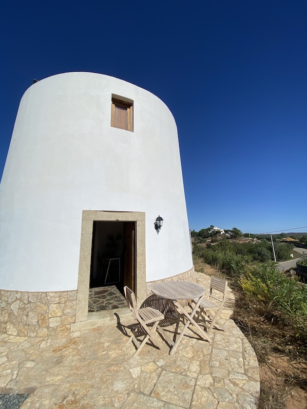 A white, rounded building with a small window and a door, a stone-paved patio with wooden furniture, and a view of a hilly landscape. - Beautiful mill and views, in the hinterland of the Algarve coast line.<br> (Alcantarilha)