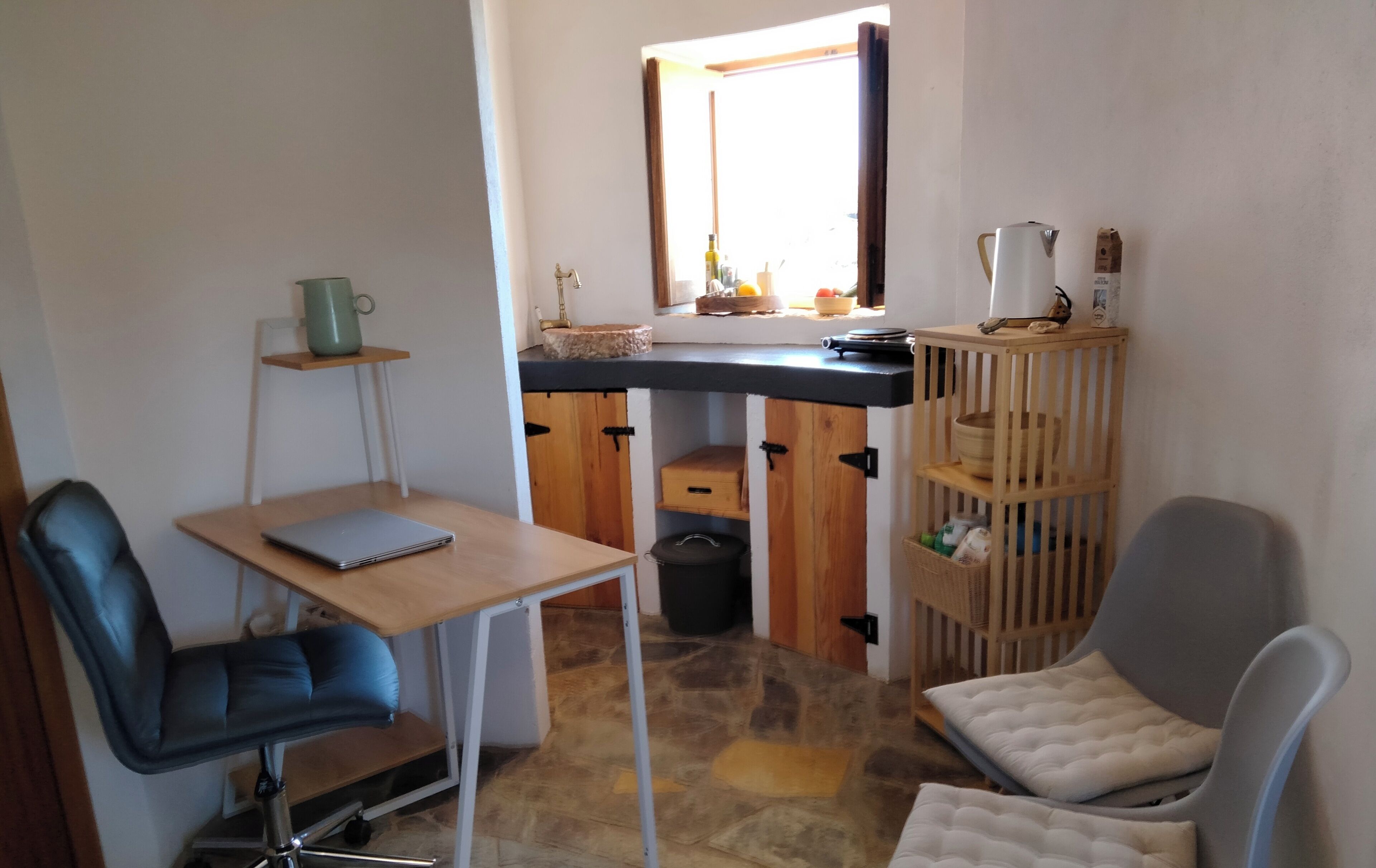 Une cuisine compacte avec un plan de travail en bois, un évier et une fenêtre au-dessus. On y trouve une petite table avec un ordinateur portable, une chaise et une étagère sur laquelle se trouve un récipient vert. Un meuble en bois avec une poubelle noire est visible.