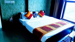 A neatly made bed with a white duvet and a colorful blanket, two pillows, a bedside table with a kettle, and a window with a view. - Mgh 114 Hotel Krishna Udaipur (Udaipur)