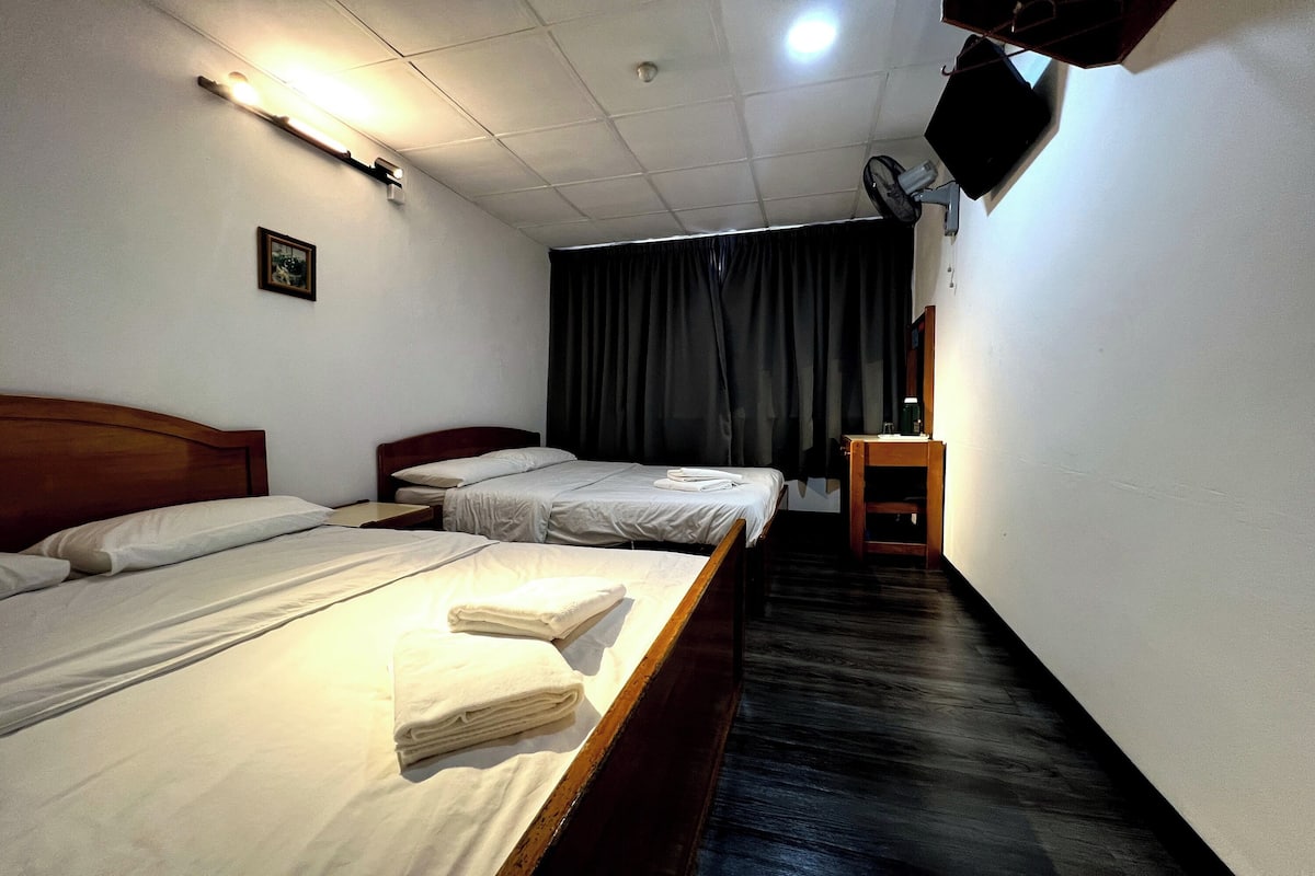Habitación de hotel con dos camas, cabecero de madera, una mesita de noche de madera, cortina oscura y televisor montado.