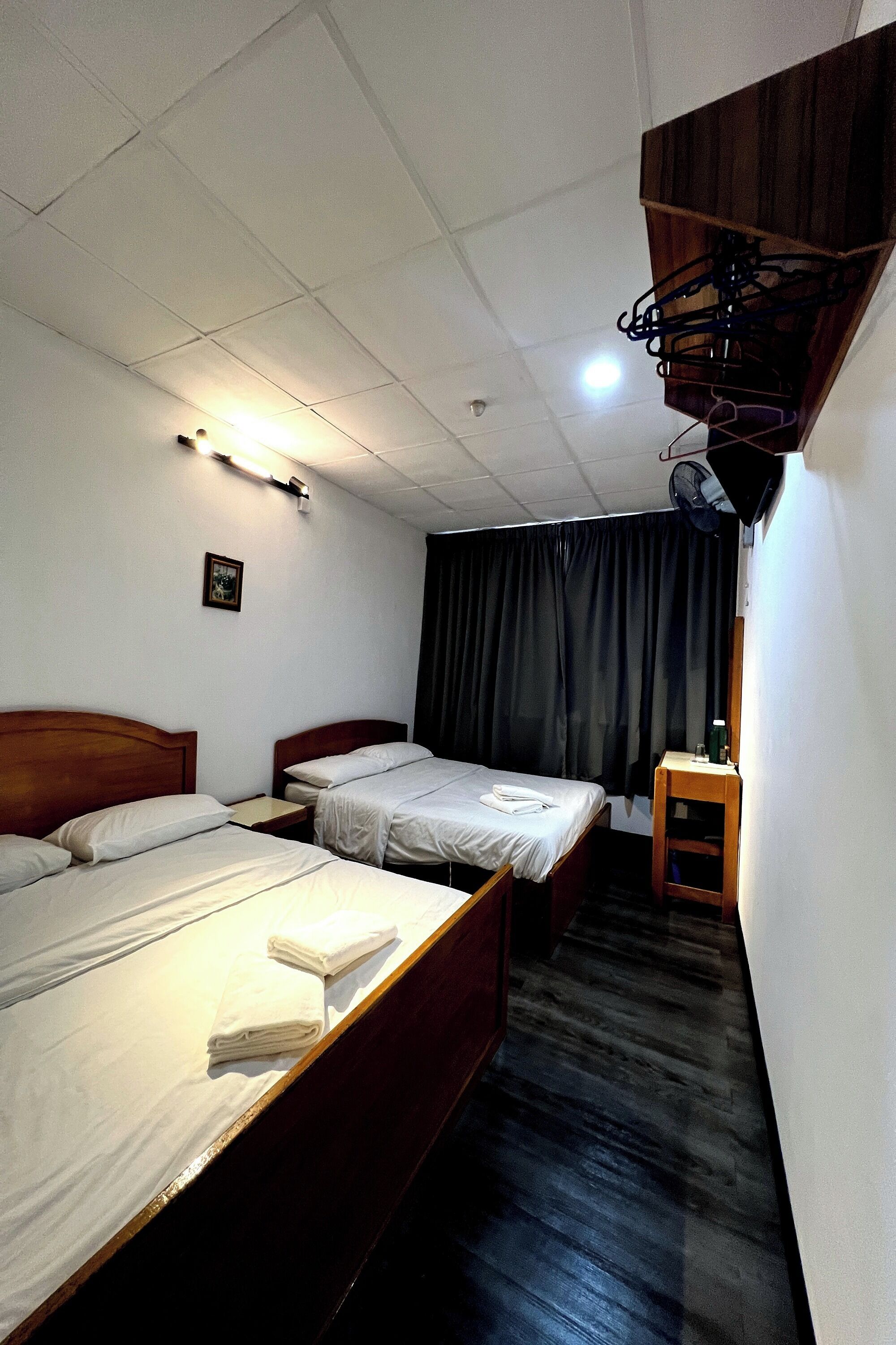 Habitación de hotel con dos camas individuales, una mesita pequeña, una silla, un ventilador de techo y una ventana con cortinas.