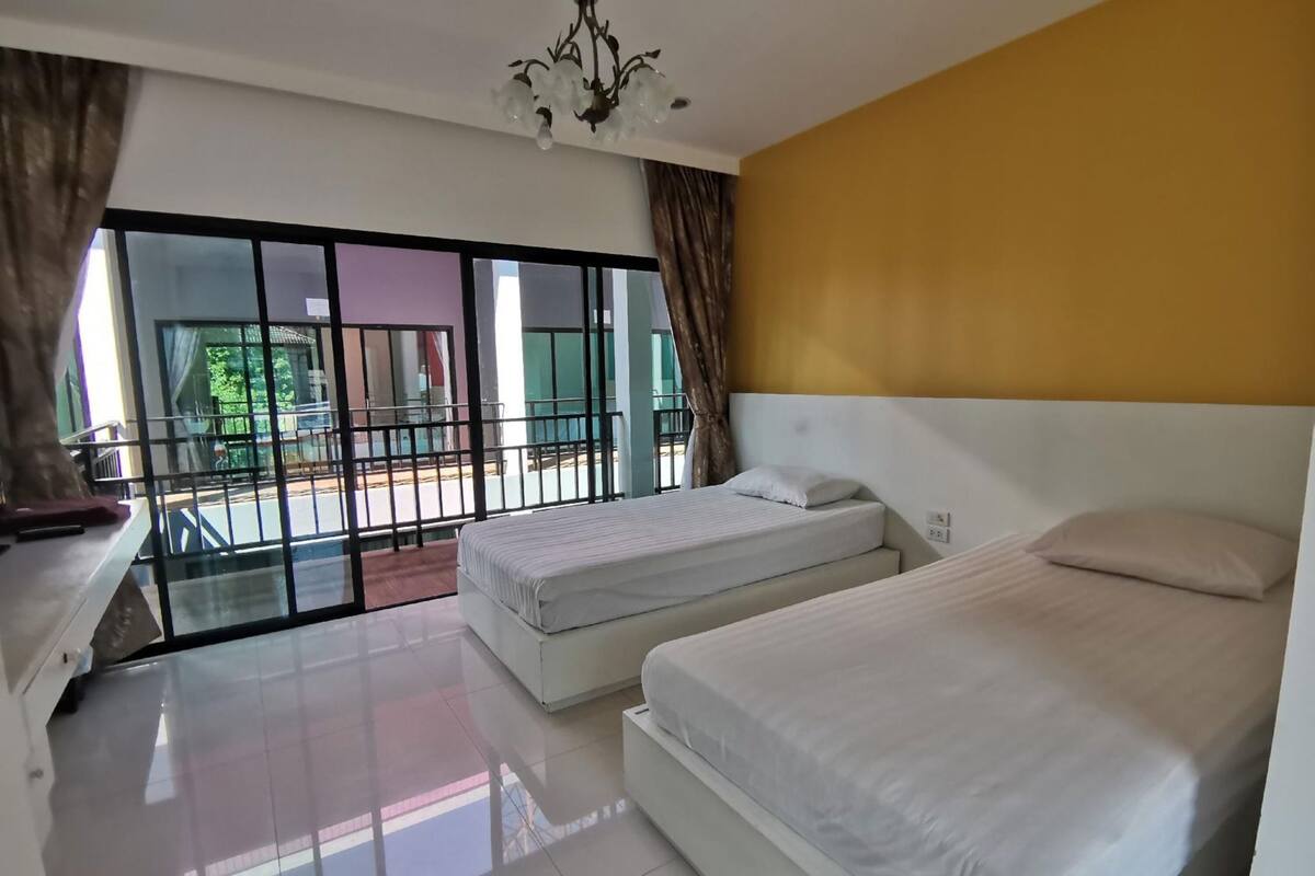 Habitación de hotel con dos camas, un candelabro, un balcón con barandilla y vistas a zonas verdes.