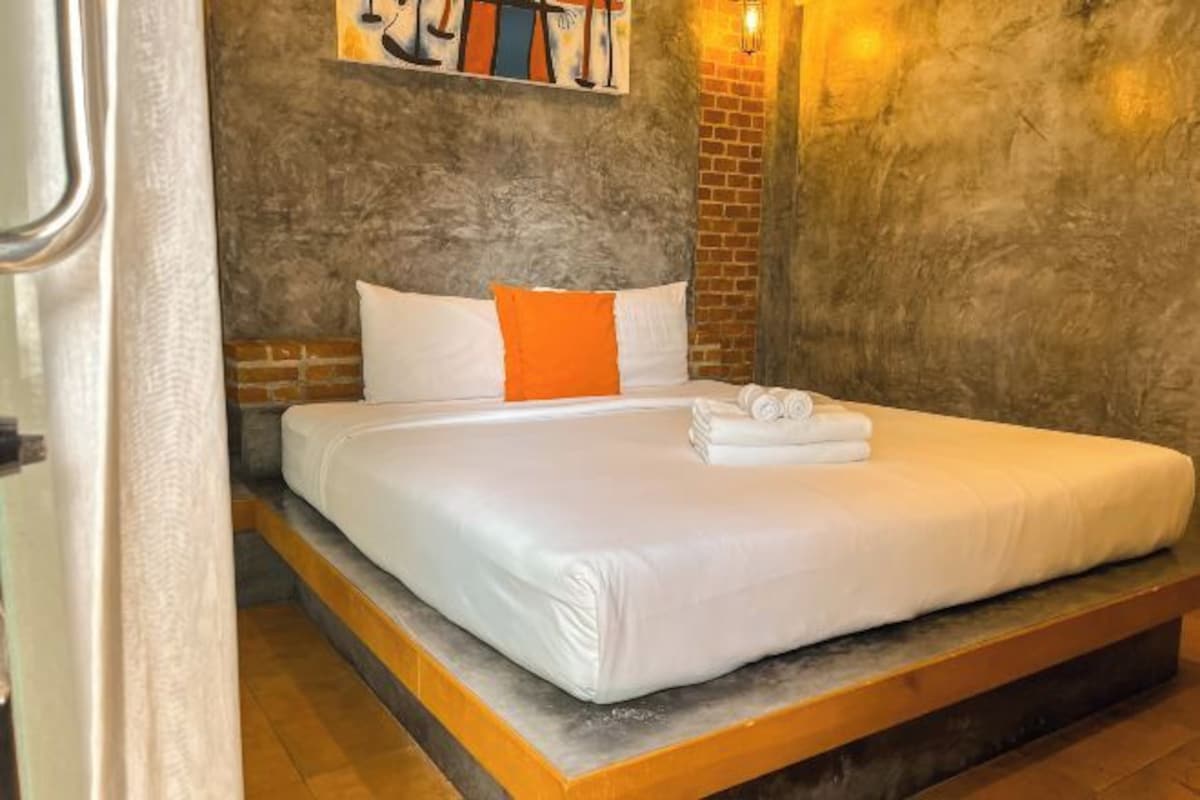 A bed with white bedding and orange pillows, a brick wall, and a painting above the bed.