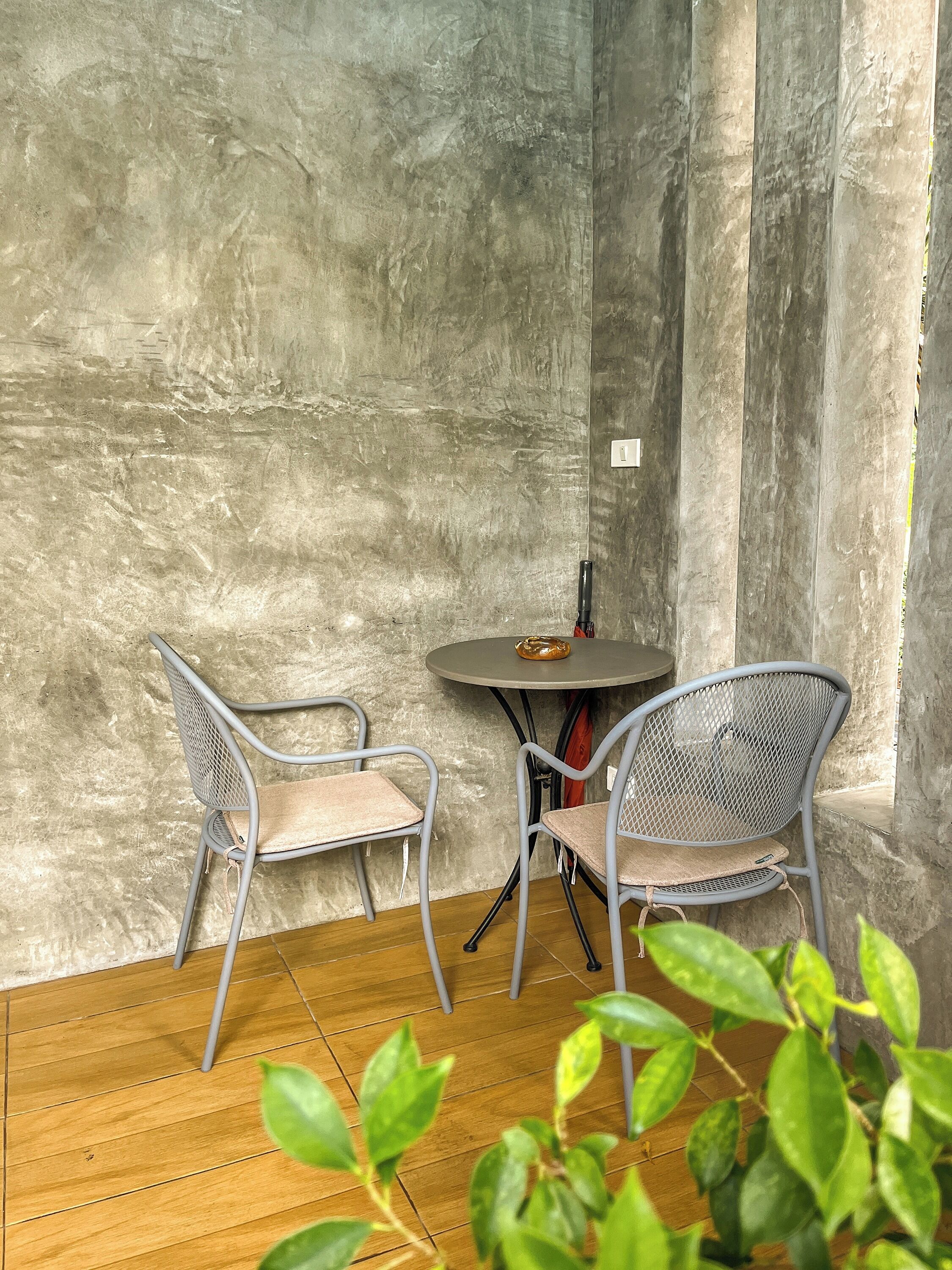 A small, enclosed patio area with two metal chairs and a round table, featuring a concrete wall and wooden flooring.
