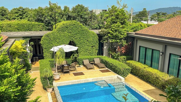 Una piscina con solárium empedrado, rodeada de exuberante vegetación y un patio con mobiliario exterior.