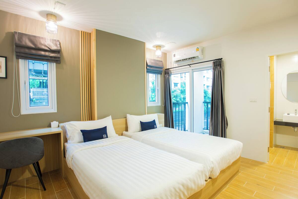 A hotel room with two beds, a desk, a chair, a bathroom with a sink, and a view of greenery outside.