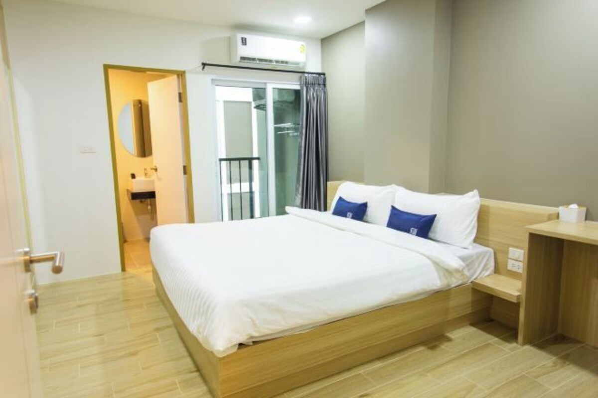 A hotel room with a wooden bed, a small bedside table, a built-in shelf, a bathroom with a glass door, and an air conditioning unit.